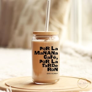 May include: A clear glass tumbler with a wooden lid and a straw. The tumbler is filled with a light brown liquid and has the text "Por La Ma&ntilde;ana Caf&eacute;, Por La Tarde Ron -Bad Bunny" printed on the side.
