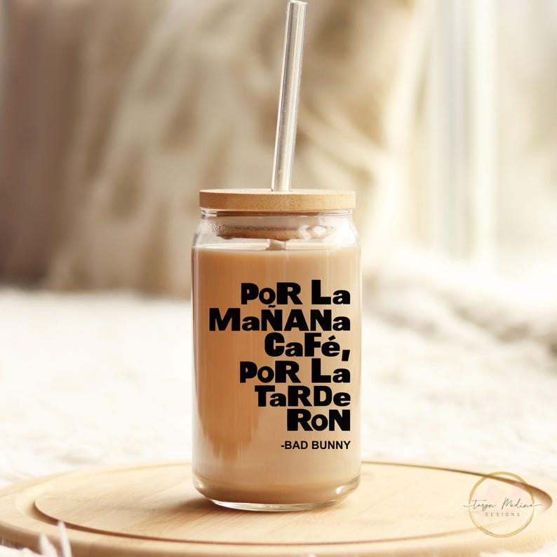 May include: A clear glass tumbler with a wooden lid and a straw. The tumbler is filled with a light brown liquid and has the text "Por La Ma&ntilde;ana Caf&eacute;, Por La Tarde Ron -Bad Bunny" printed on the side.