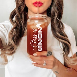 May include: A clear plastic tumbler with a brown beverage inside. The tumbler has a white design that says "Class of 2025" with a graduation cap icon.