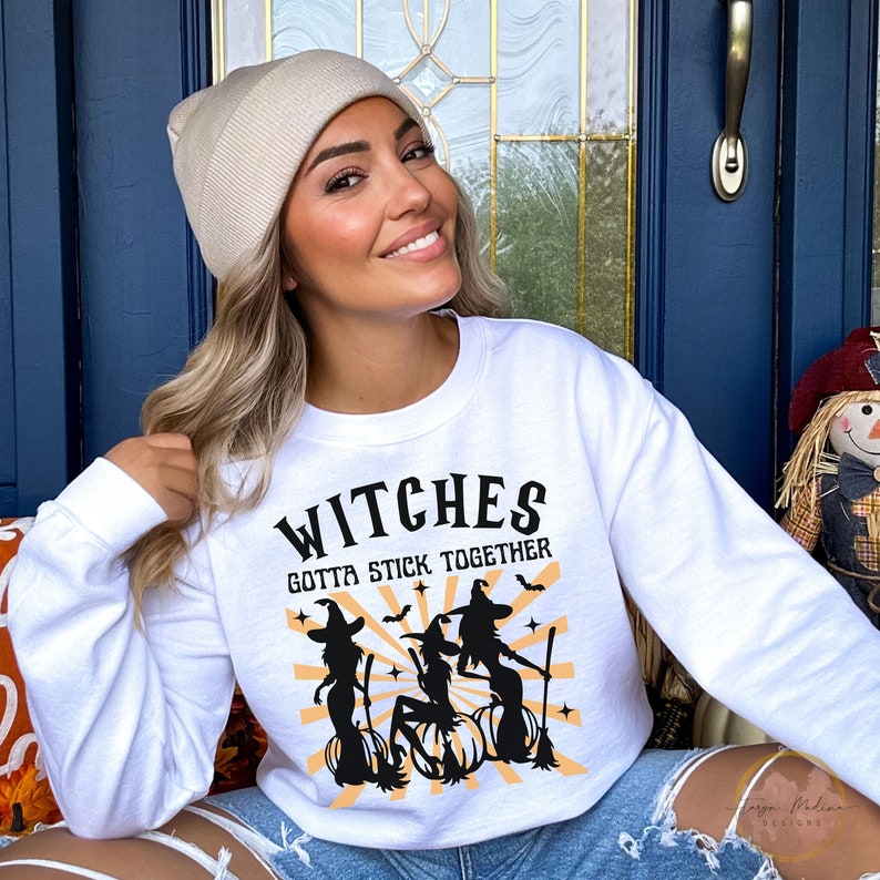 Puede incluir: Sudadera blanca con un gr&aacute;fico negro de tres brujas en escobas con el texto "Witches Gotta Stick Together".