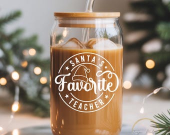 Puede incluir: Un vaso transparente con tapa de bambú y pajita de cristal, lleno de una bebida de color marrón claro y hielo. La parte frontal presenta un gráfico circular blanco con un icono de gorro de Papá Noel y el texto "SANTA'S Favorite TEACHER".