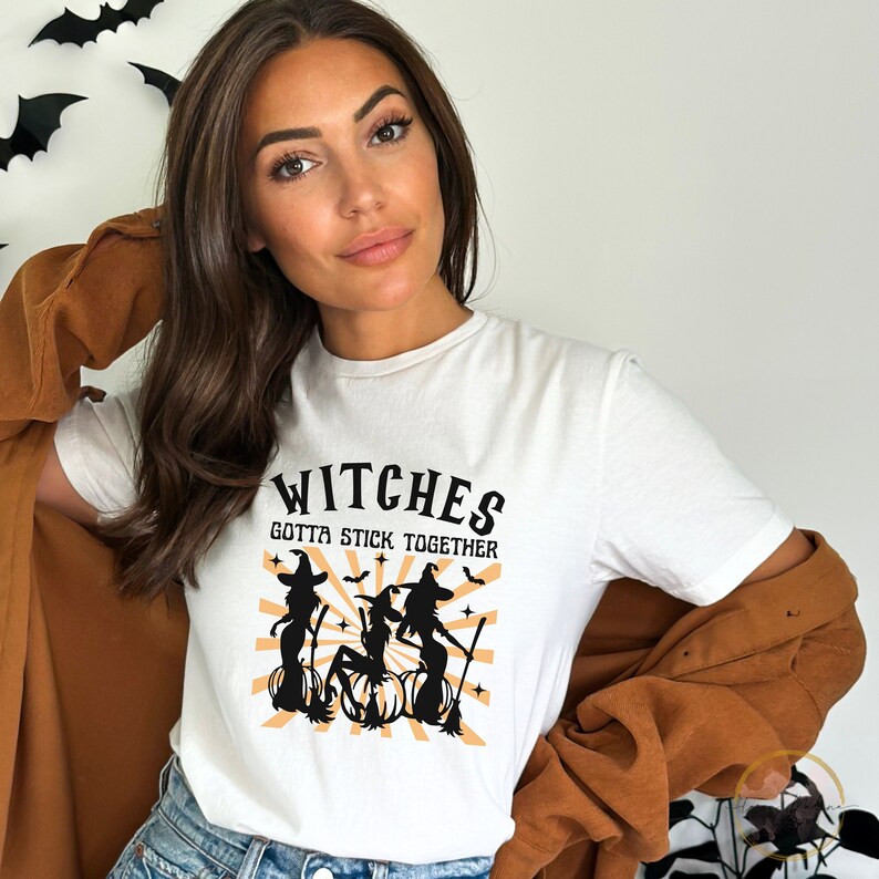 Puede incluir: Camiseta blanca con un gr&aacute;fico negro de tres brujas sobre calabazas con el texto "Witches Gotta Stick Together".