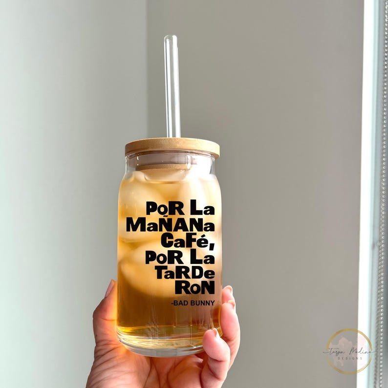 May include: A clear glass jar with a wooden lid and a straw. The jar is filled with a light brown liquid and ice. The jar has a black and white sticker with the text "Por La Ma&ntilde;ana Caf&eacute;, Por La Tarde Ron - Bad Bunny".