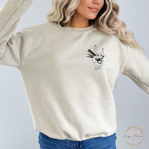 Peut inclure: Un sweat-shirt beige clair avec un motif noir d'une main tenant des ciseaux et des peignes dans un geste de rock and roll. Le motif est sur le c&ocirc;t&eacute; gauche de la poitrine.