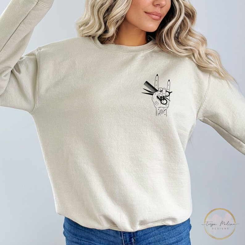 Peut inclure: Un sweat-shirt beige clair avec un motif noir d'une main tenant des ciseaux et des peignes dans un geste de rock and roll. Le motif est sur le c&ocirc;t&eacute; gauche de la poitrine.