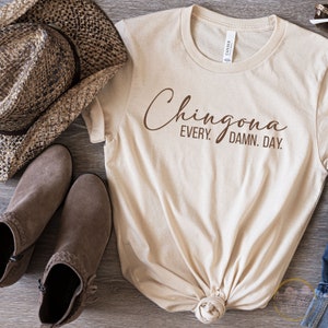 May include: A beige t-shirt with the text "Chingona Every. Damn. Day." printed in brown on the front. The shirt is knotted at the bottom and is laying on a wooden surface with a brown cowboy hat and brown boots.