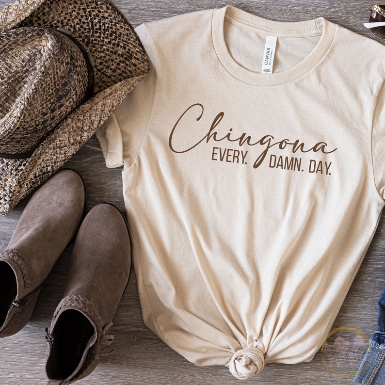 May include: A beige t-shirt with the text "Chingona Every. Damn. Day." printed in brown on the front. The shirt is knotted at the bottom and is laying on a wooden surface with a brown cowboy hat and brown boots.
