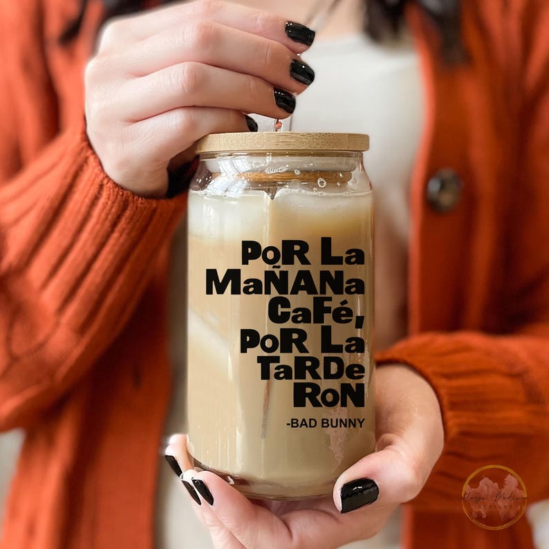 May include: A glass jar with a wooden lid filled with a light brown beverage. The jar has black text that reads "Por La Ma&ntilde;ana Caf&eacute;, Por La Tarde Ron - Bad Bunny".