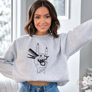 Peut inclure: Un sweat-shirt gris avec un motif noir d'une main tenant des ciseaux et un peigne. La main fait un geste de rock and roll. Le motif est entour&eacute; de petits d&eacute;tails comme des &eacute;toiles, des lunes et une rose.