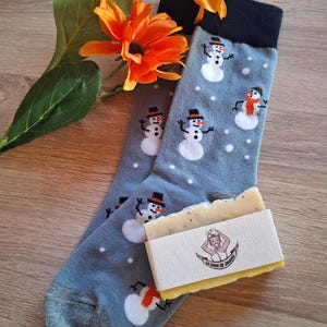 Könnte beinhalten: Graue Socken mit Schneemannmuster, die Schneemänner mit schwarzen Hüten und orangefarbenen Schals zeigen. Eine Seife mit Logo ist ebenfalls vorhanden, zusammen mit orangefarbenen Blumen.