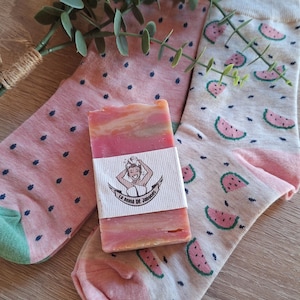 May include: A bar of pink and orange soap with a label featuring a woman's illustration, placed between two pairs of socks. One pair of socks is pink with blue dots, and the other is light gray with watermelon slices.