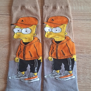 Può includere: Un paio di calzini con un personaggio dei cartoni animati con una felpa con cappuccio arancione, un berretto e pantaloni neri. I calzini hanno una combinazione di colori grigio e marrone, con il volto e l'abbigliamento del personaggio. La parola "PALACE" è stampata sul lato dei pantaloni.
