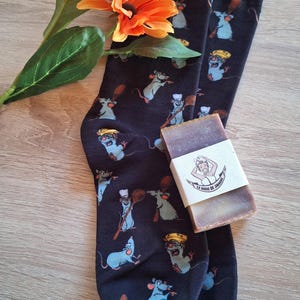 Könnte beinhalten: Schwarze Socken mit einem Muster aus Cartoon-Ratten mit Kochmützen und Kochutensilien. Ein Seifenstück mit weißem Etikett liegt auf den Socken. Orangefarbene Blumen und grüne Blätter sind im Hintergrund.