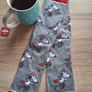 以下が含まれることがあります： 赤いつま先と袖口が付いたグレーの靴下で、スケートボードをするスヌーピーの繰り返し模様が特徴です。白い水玉模様の薄い青色のマグカップと紅茶のカップも画像にあります。