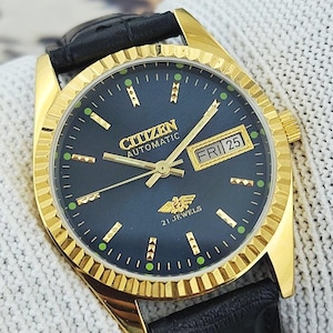 May include: A gold Citizen automatic wristwatch with a dark blue face and black leather strap. The watch face shows the day and date, with gold hands and markers. The watch is marked "21 Jewels".