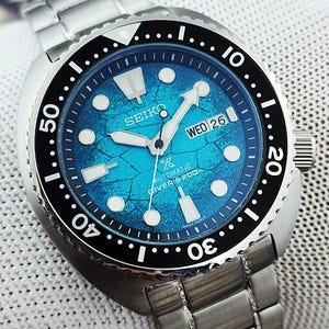 May include: A stainless steel Seiko automatic diver's watch with a blue dial. The watch features a black rotating bezel, luminous hands and markers, and a date display. The watch band is made of stainless steel.