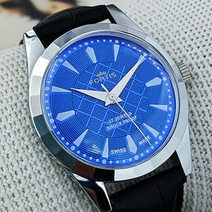 May include: A silver wristwatch with a bright blue face displaying a geometric design. The watch has white hands and a black leather strap. The face includes the text "FORTIS", "17 JEWELS SHOCK PROOF", and "SWISS MADE".