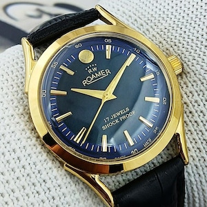 Puede incluir: Un reloj de pulsera dorado con esfera azul oscuro y agujas e indicadores dorados. La correa es de cuero negro. La esfera muestra "ROAMER" y "17 JEWELS SHOCK PROOF". El fondo es blanco y texturizado.