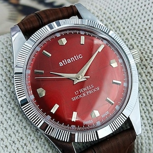 Puede incluir: Un reloj de pulsera Atlantic vintage con esfera roja y correa de cuero marrón. El reloj presenta agujas e indicadores plateados, y la esfera indica "atlantic", "17 jewels" y "shock proof". El reloj tiene un bisel plateado y una correa de cuero marrón texturizada.