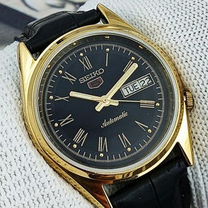 Könnte beinhalten: Eine goldfarbene Seiko Automatik-Armbanduhr mit schwarzem Zifferblatt, goldenen Zeigern und römischen Ziffern. Die Uhr hat eine Tages- und Datumsanzeige und ein schwarzes Lederarmband.