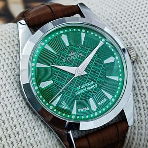 May include: A silver wristwatch with a green patterned face and white hands. The watch has a brown leather strap. The watch face includes the text "17 JEWELS SHOCK PROOF SWISS MADE".