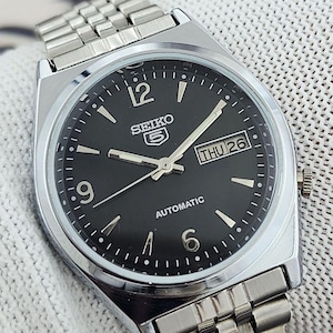 May include: A silver-toned Seiko 5 automatic watch with a black dial. The watch features silver hands and markers, a day-date display, and a stainless steel band. The watch face displays the day as "THU 26".