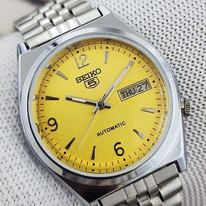 May include: A silver-toned Seiko 5 automatic watch with a bright yellow face. The watch features black hour markers, a day-date display, and a silver metal band. The watch face displays the day as "THU 27".