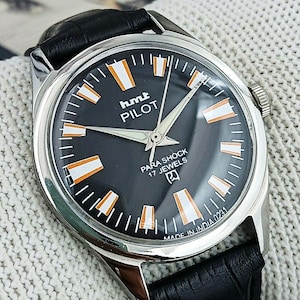 Może przedstawiać: Zegarek HMT Pilot w stylu vintage z czarną tarczą, srebrną kopertą i czarnym skórzanym paskiem. Tarcza zegarka posiada białe i pomarańczowe znaczniki, srebrne wskazówki oraz napis "hmt PILOT PARA SHOCK 17 JEWELS". Zegarek oznaczony napisem "MADE IN INDIA 0231".