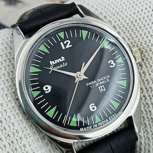 May include: A vintage HMT Janata wristwatch with a black dial and silver case. The watch features green accents, white numerals, and the text "PARA SHOCK 17 JEWELS". It has a black leather strap and a silver crown.