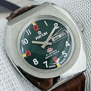 Puede incluir: Un reloj Ricoh vintage con esfera verde oscuro, caja plateada y correa de cuero marrón. La esfera muestra números blancos, detalles en rojo y el día y la fecha. El reloj está marcado "21 Jewels Shock Proof" y "Made in Japan."