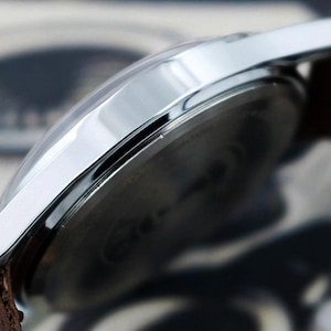 May include: Close-up of a silver wristwatch with a brown leather strap. The watch has a clear, curved crystal and a shiny, metallic bezel. The watch face is dark, and the strap is textured.