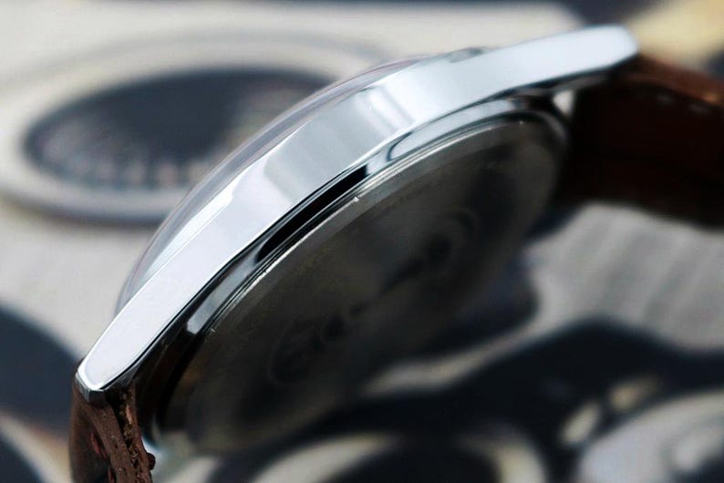 May include: Close-up of a silver wristwatch with a brown leather strap. The watch has a clear, curved crystal and a shiny, metallic bezel. The watch face is dark, and the strap is textured.