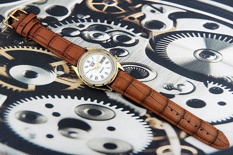 May include: A gold-toned wristwatch with a white face, Roman numeral hour markers, and a brown leather band. The watch features a day and date display. The band has a crocodile pattern. The watch is set against a background of gears.