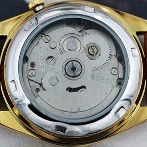 May include: Close-up of a gold-toned wristwatch with a brown leather band. The watch's back is visible, showcasing the intricate internal mechanisms, including gears and the Seiko brand name. The watch has a silver bezel.