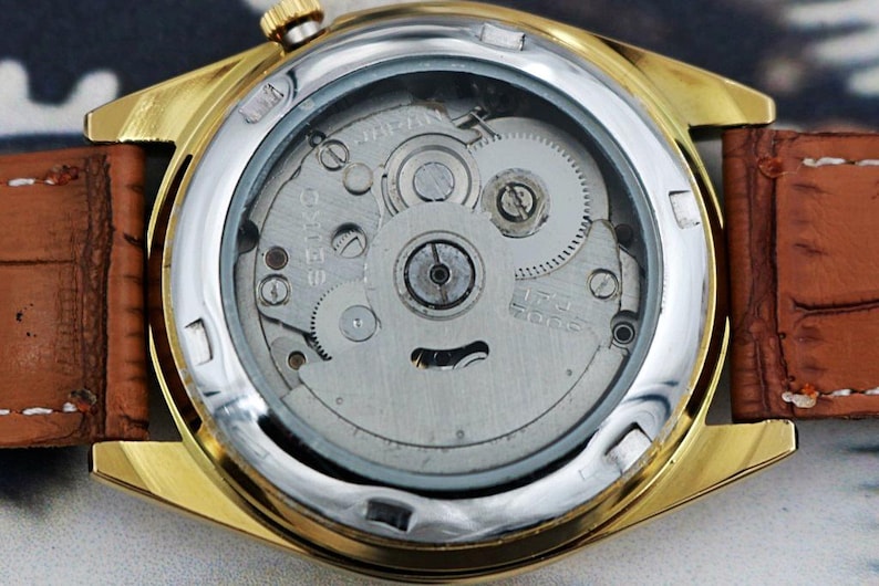 May include: Close-up of a gold-toned wristwatch with a brown leather band. The watch's back is visible, showcasing the intricate internal mechanisms, including gears and the Seiko brand name. The watch has a silver bezel.