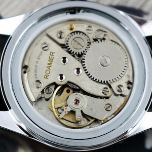 May include: Close-up of a vintage Roamer watch, showcasing the intricate mechanical movement. The watch features a silver case, brown leather strap, and visible gears, springs, and the brand name "ROAMER". The watch's internal components are visible through the clear back.
