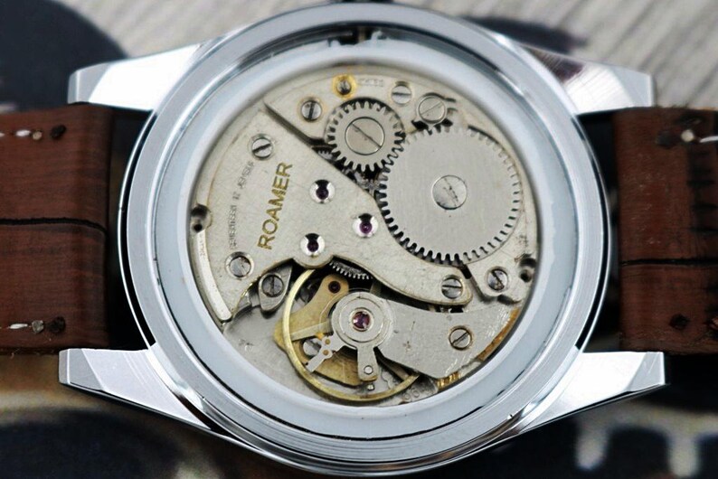 May include: Close-up of a vintage Roamer watch, showcasing the intricate mechanical movement. The watch features a silver case, brown leather strap, and visible gears, springs, and the brand name "ROAMER". The watch's internal components are visible through the clear back.