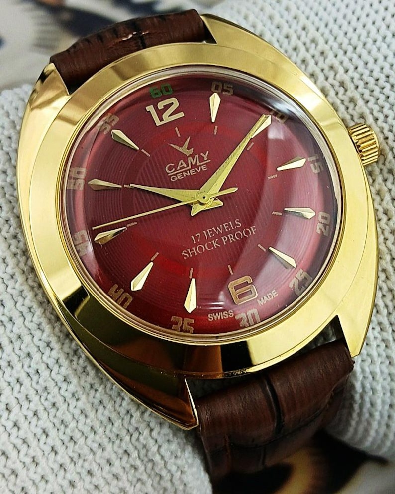 K&ouml;nnte beinhalten: Eine goldfarbene Armbanduhr mit burgunderfarbenem Zifferblatt und braunem Lederarmband. Das Zifferblatt der Uhr zeigt goldene Zeiger und Markierungen, mit dem Markennamen "CAMY GENEVE" und dem Text "17 JEWELS SHOCK PROOF". Die Uhr hat ein klassisches Design.