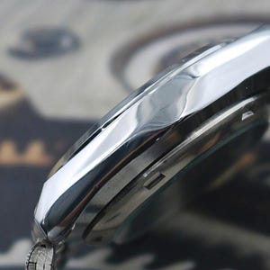 May include: Close-up of a silver wristwatch. The watch has a polished, reflective bezel and a silver band. The watch face is partially visible, with a dark background. The watch appears to be a classic design.