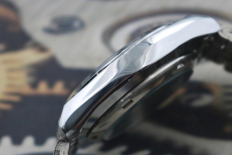 May include: Close-up of a silver wristwatch. The watch has a polished, reflective bezel and a silver band. The watch face is partially visible, with a dark background. The watch appears to be a classic design.