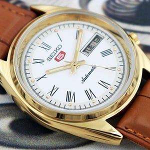 May include: A gold-toned Seiko 5 automatic watch with a white face, Roman numeral hour markers, and a day-date display. The watch has gold hands and a brown leather strap.