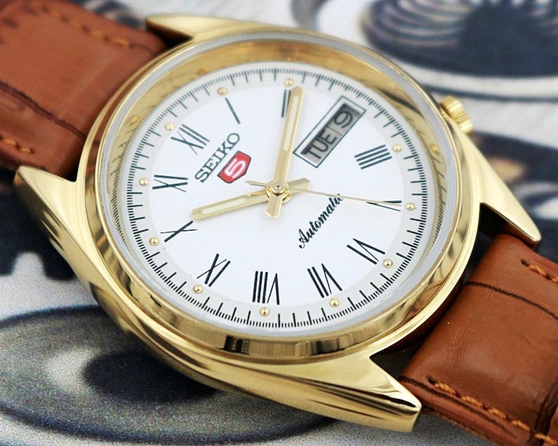 May include: A gold-toned Seiko 5 automatic watch with a white face, Roman numeral hour markers, and a day-date display. The watch has gold hands and a brown leather strap.