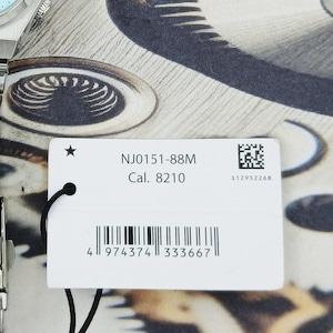 May include: Close-up of a silver wristwatch with a light blue face and a stainless steel band. A white tag with the text "NJ0151-88M Cal. 8210" and a barcode is attached to the watch.