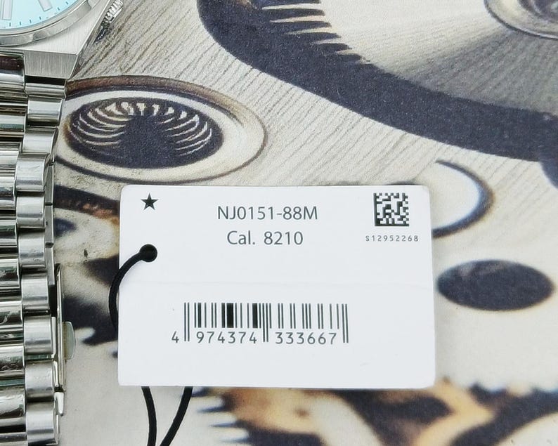 May include: Close-up of a silver wristwatch with a light blue face and a stainless steel band. A white tag with the text "NJ0151-88M Cal. 8210" and a barcode is attached to the watch.