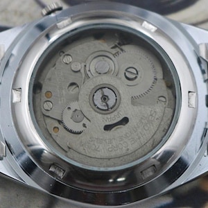 May include: Close-up of the back of a silver-toned wristwatch, showcasing the intricate internal mechanism. The watch face is transparent, revealing gears, springs, and other components. The watch band is made of linked metal segments.