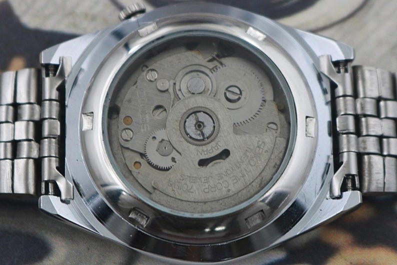 May include: Close-up of the back of a silver-toned wristwatch, showcasing the intricate internal mechanism. The watch face is transparent, revealing gears, springs, and other components. The watch band is made of linked metal segments.