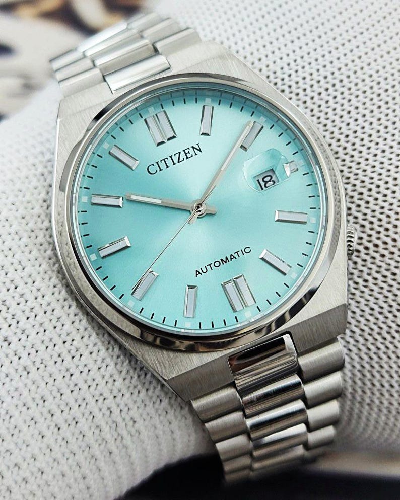 May include: A silver Citizen automatic watch with a light blue face and a date display. The watch has silver hands and markers, and a silver link band. The watch is displayed on a white textured surface.