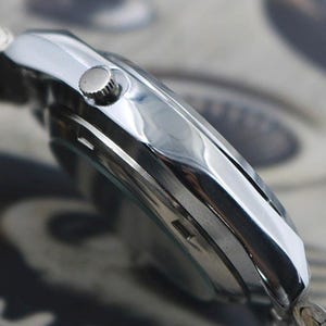 May include: Close-up of a silver wristwatch with a polished metal band. The watch face is not visible, but the side profile shows a round crown and a sleek, reflective surface. The watch appears to be new and unworn.