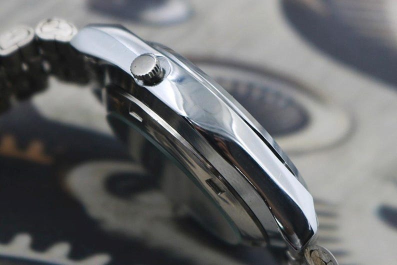 May include: Close-up of a silver wristwatch with a polished metal band. The watch face is not visible, but the side profile shows a round crown and a sleek, reflective surface. The watch appears to be new and unworn.