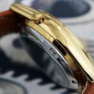 May include: Close-up of a gold-toned wristwatch with a brown leather band. The watch has a gold bezel and a silver-colored face. The band is a light brown color.